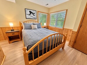 Quarto com cama de madeira, mesa de cabeceira, abajur e janela com persianas.