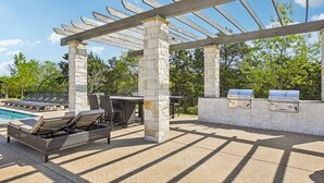 Een terras buiten met een pergola, barbecueplaatsen, een zwembad en lounge stoelen.