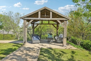 A wooden gazebo with two wicker chairs and a small table, surrounded by a fenced garden.