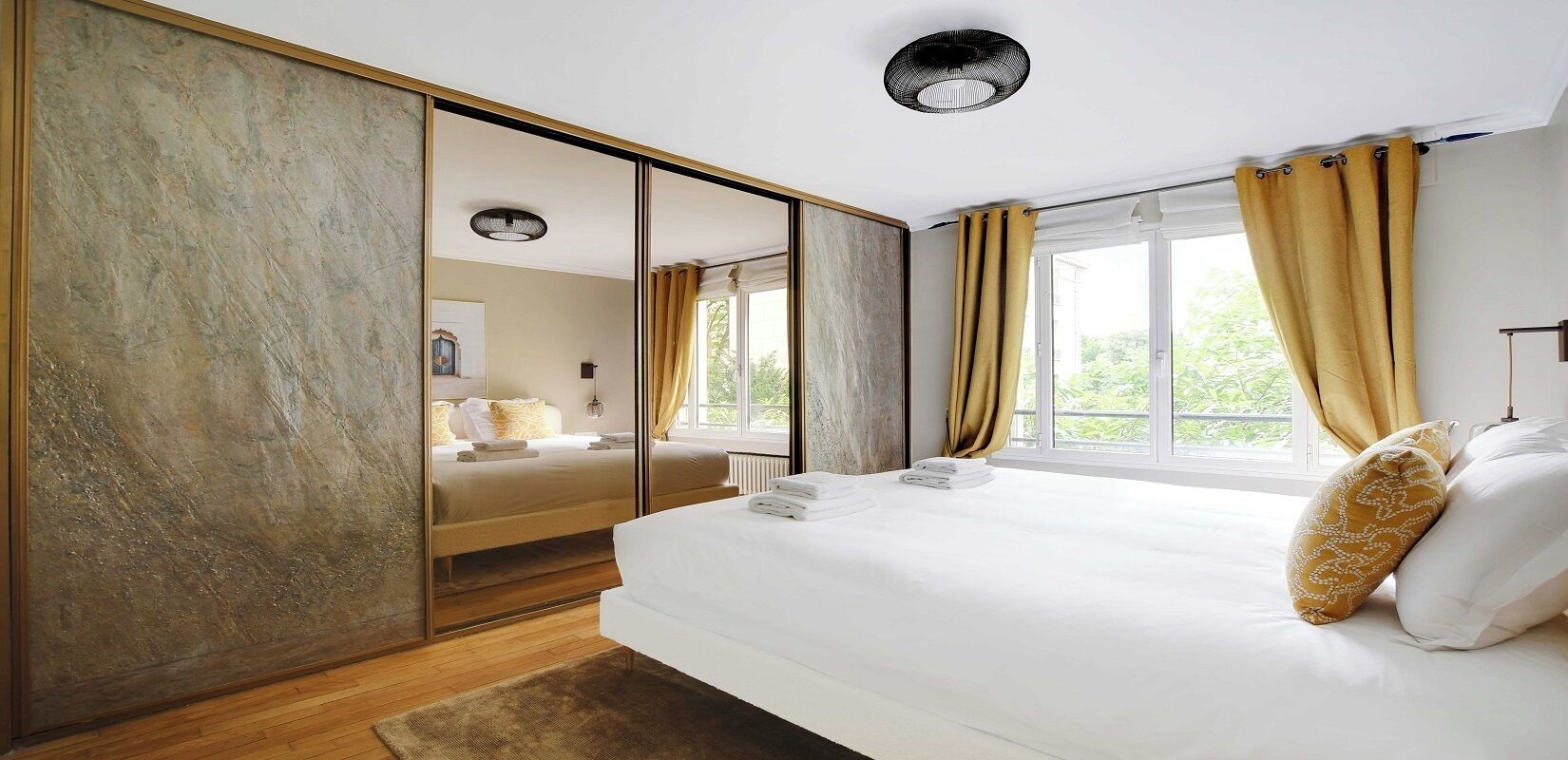 A modern bedroom with a large bed, a mirror, and a view of greenery outside.