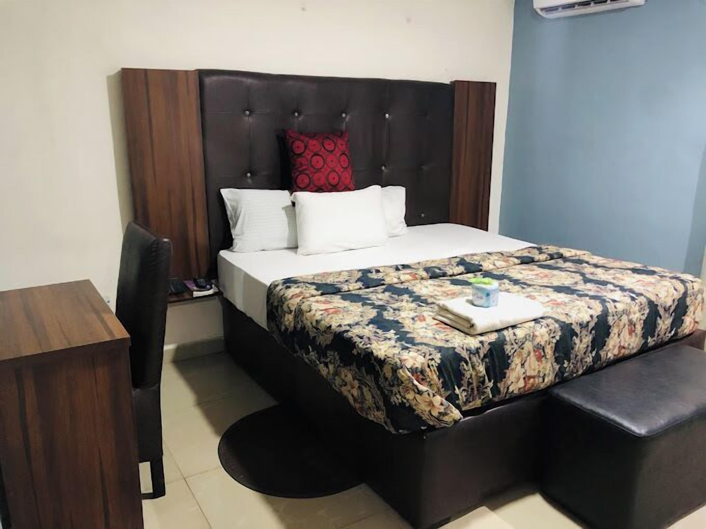 Una cama bien hecha con una colcha estampada de flores, un cojín con estampado rojo y almohadas blancas. Hay una mesita de noche con una lámpara y una silla.