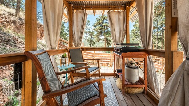 A wooden deck with two chairs and a table, a grill, and a propane tank. - Glamping Under the Stars | Firepit, BBQ & Views (Woodland Park)