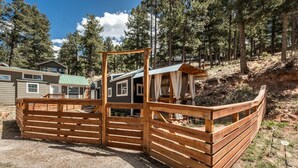 A wooden fence enclosure with a metal gate, surrounded by a forested area and a small cabin. - Glamping Under the Stars | Firepit, BBQ & Views (Woodland Park)