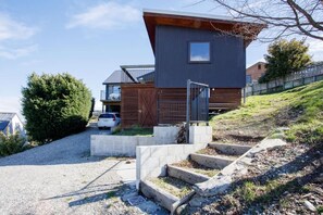 A modern two-story house with a sloped roof, a garage, and a driveway leading to a gated entrance. - Grandview Vista - Lake Hawea (Lake Hawea)