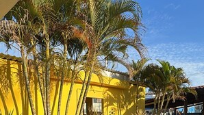 A yellow building with a tiled patio area, surrounded by various plants and palm trees. - Pousada Vila do Sol (Ipojuca)