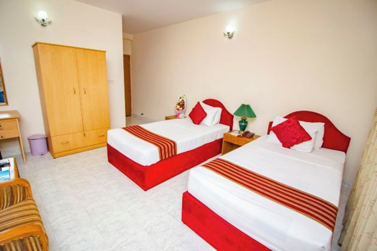 A hotel room with two beds, each with red and white bedding, a wooden wardrobe, a desk with a lamp, and a trash bin.