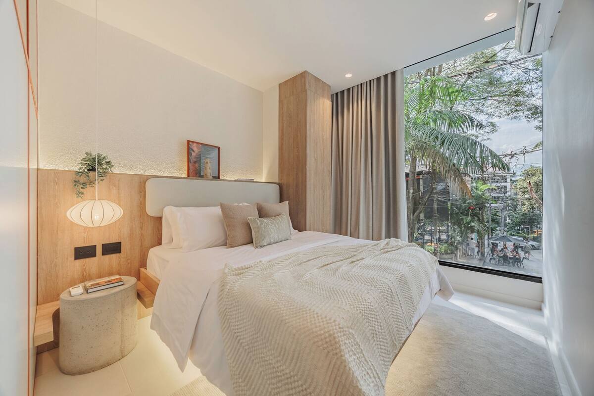 A modern bedroom with a large window, a bed with white linens, a bedside table with a lamp, and a view of the outdoors.