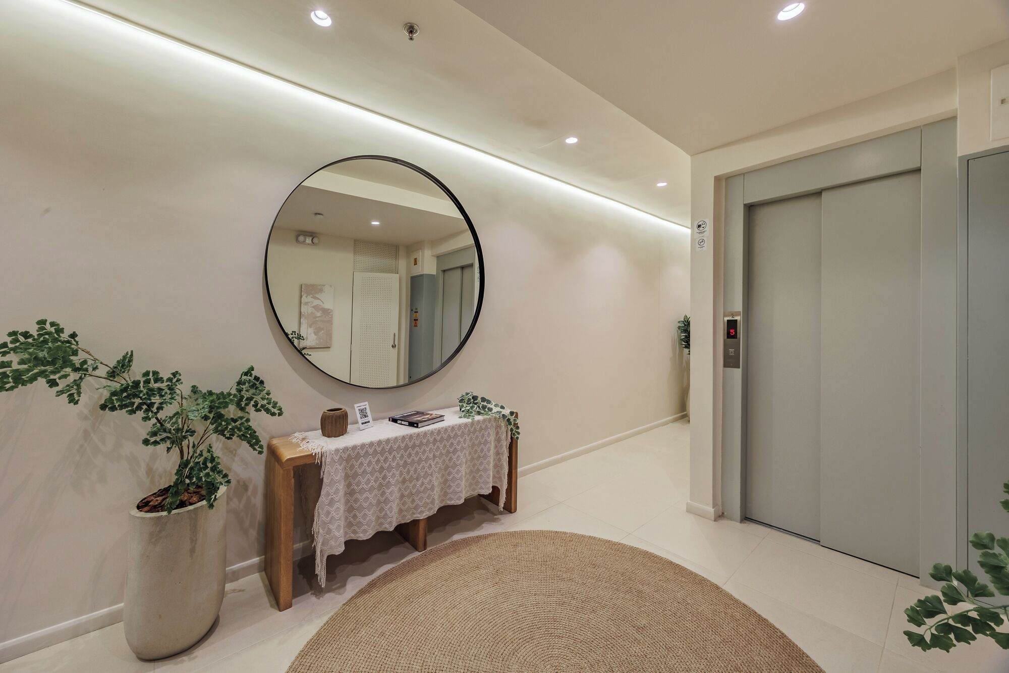 Un couloir avec un grand miroir rond, une petite table ornée d’un vase et de quelques livres, une plante en pot et un tapis à motifs.