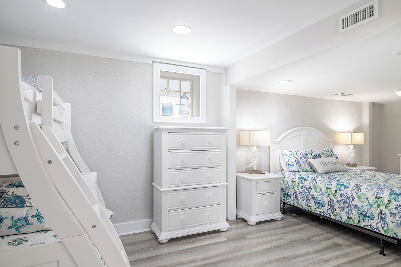 A bedroom with a white dresser, a bed with floral bedding, a nightstand with lamps, and a window with a view.
