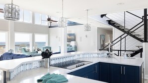 Une cuisine moderne dotée d’un grand îlot central, de luminaires suspendus et offrant une vue sur l’océan.