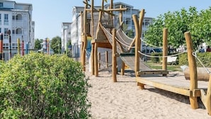 A wooden play structure with a rope bridge and slide, surrounded by sand and greenery, with buildings in the background. - Ferienwohnungen im Seepark Sellin (Sellin)