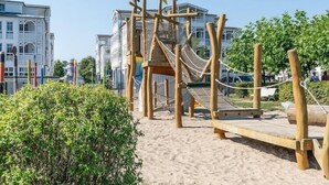 A wooden play structure with a rope bridge and slide, surrounded by sand and greenery, with buildings in the background. - Ferienwohnungen im Seepark Sellin (Sellin)