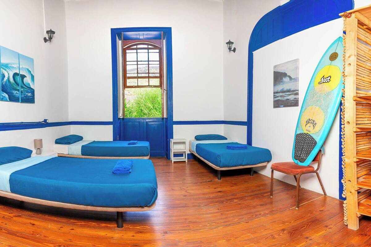 Quarto com três camas, uma prateleira de madeira, uma prancha de surf e uma janela com vista para o verde.