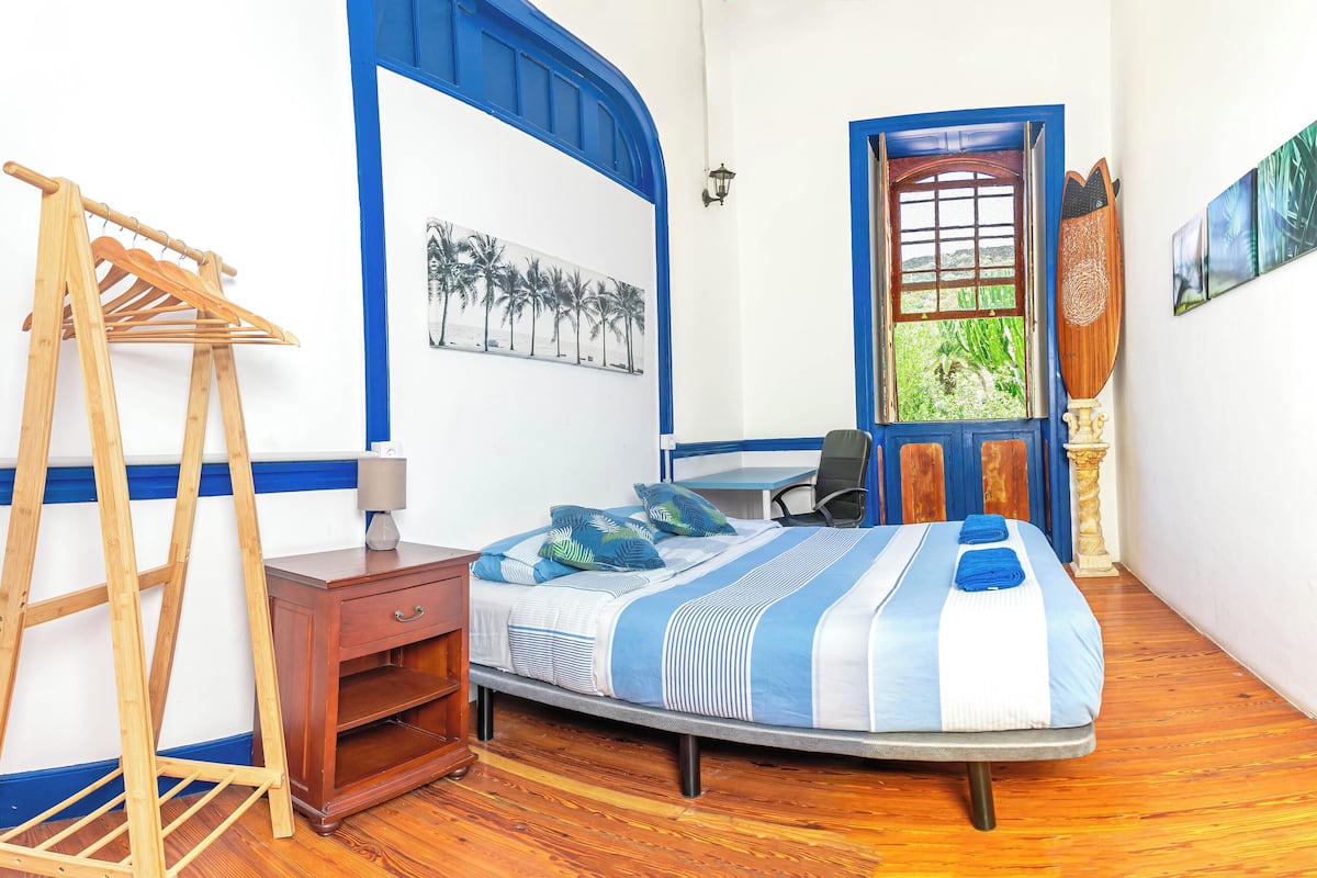 Um quarto com cama, escrivaninha, cadeira, escada de madeira e janela com vista para o verde.
