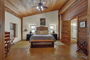 Un dormitorio con techo de madera, una cama con cabecera, dos mesitas de noche con lámparas, un banco y una puerta.