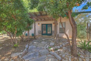 A rustic building with a stone pathway leading to a green door, surrounded by trees and a pergola.