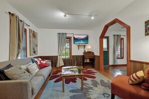 A living room with a sofa, a coffee table, and a bookshelf. There is a painting on the wall and a window with curtains. - Nandina Cottage Hot-Tub Four Minutes to Marktplatz (Fredericksburg)
