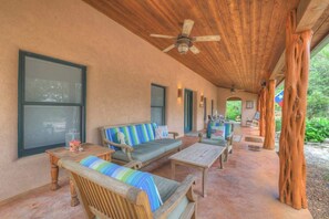 An outdoor covered patio with wooden furniture, a ceiling fan, and a view of the surrounding greenery.