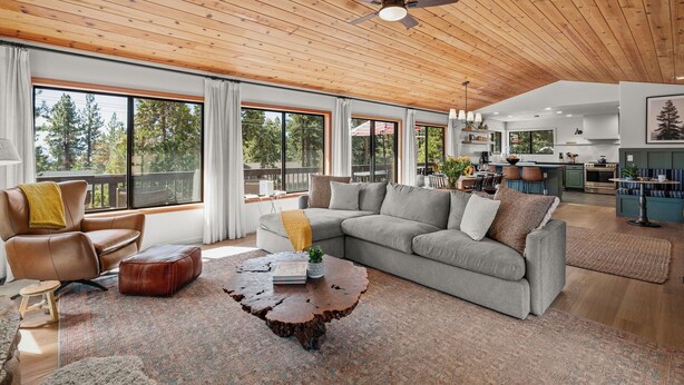Un vaste espace de vie doté d’un plafond en bois, de grandes fenêtres, d’un canapé gris, d’un fauteuil en cuir marron et d’une table basse en bois.