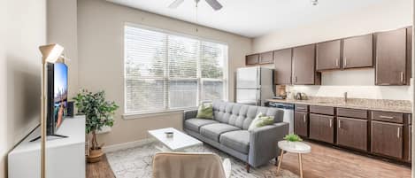 Ein modernes Wohnzimmer mit grauem Sofa, einem Couchtisch und einem Fernseher. Es gibt einen Deckenventilator, ein Fenster mit Jalousien und einen Küchenbereich mit dunklen Schränken und einem Kühlschrank.