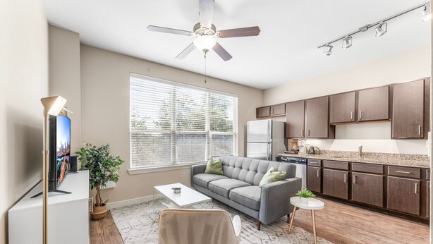 A modern living room with a grey sofa, a coffee table, and a TV. There's a ceiling fan, a window with blinds, and a kitchen area with dark cabinets and a refrigerator. - Fun Soco 1BR King Suite w Gympickleballlaundry (Austin)