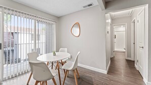 Un comedor moderno con una mesa redonda y cuatro sillas, un ventanal con persianas y un pasillo que conduce a otra habitación.