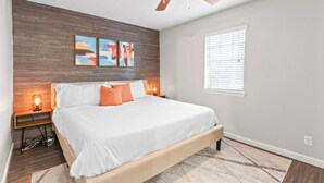 Un dormitorio con cama, paneles de madera, ventilador de techo, obras de arte, lámparas de noche y una ventana con persianas.