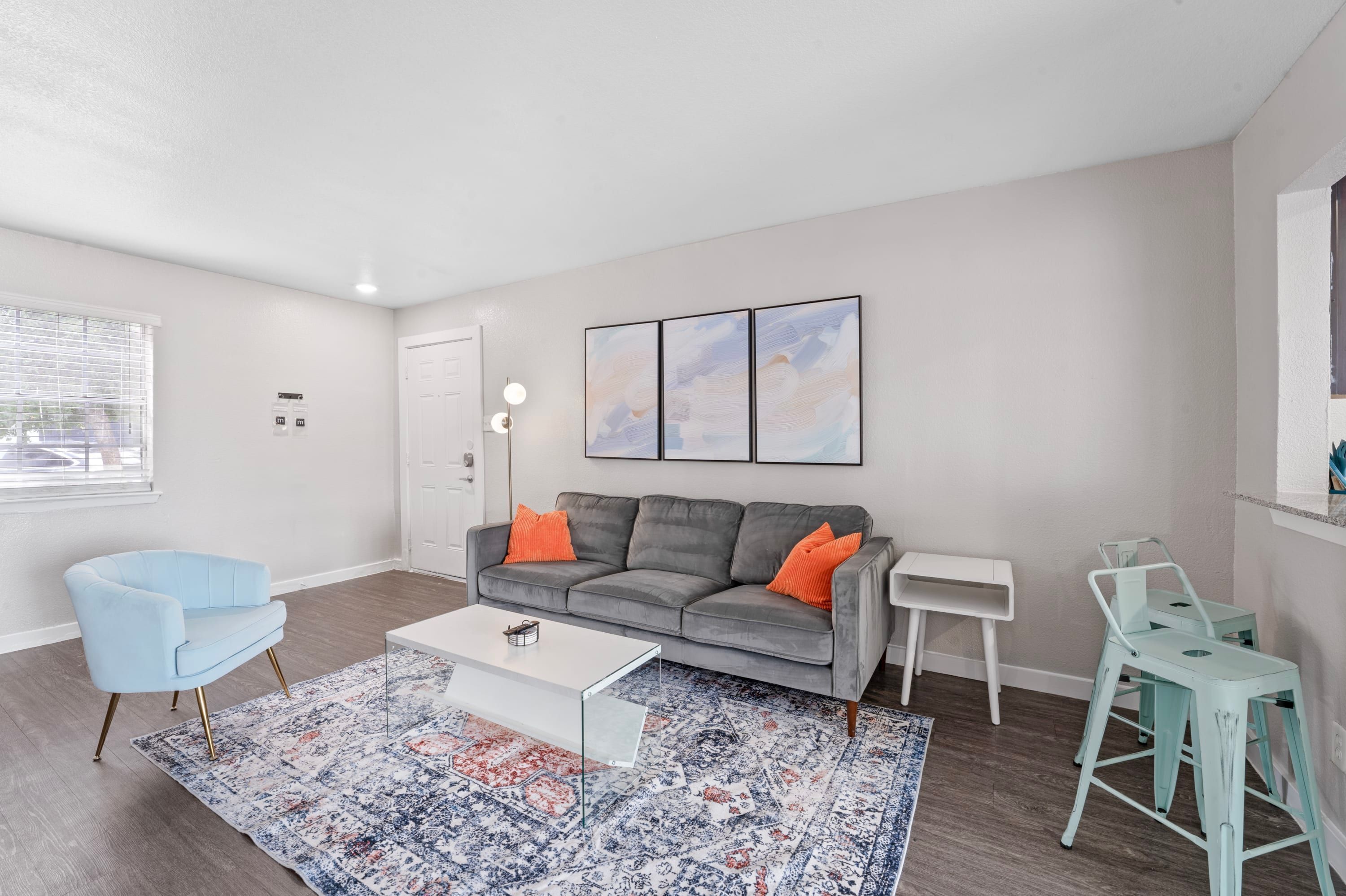 A modern living room with a grey sofa, blue armchair, and a coffee table. There are two small white side tables and a teal stool.