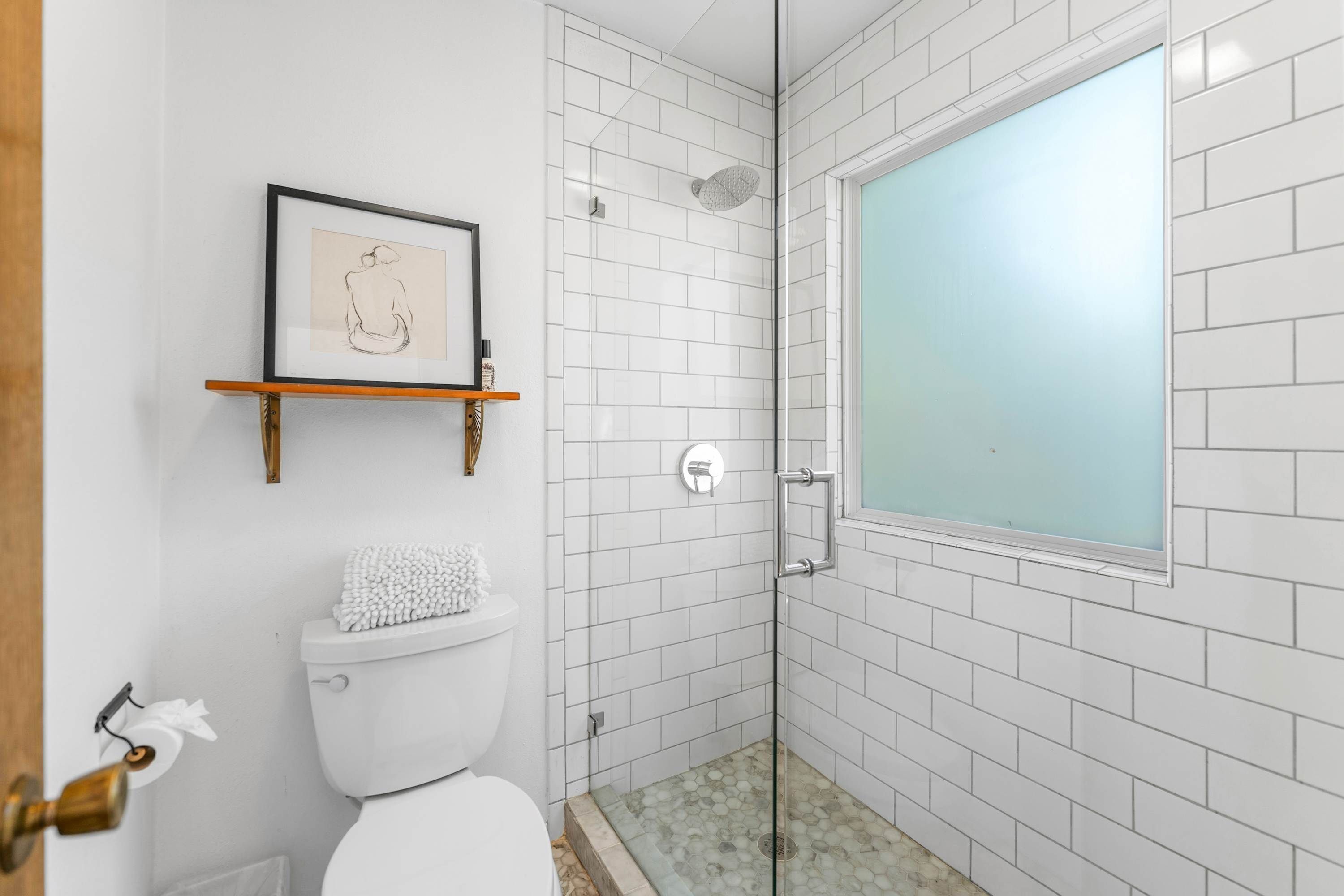 A bathroom with a white toilet, a framed picture on a shelf, a shower with a frosted glass door, and white tiled walls.