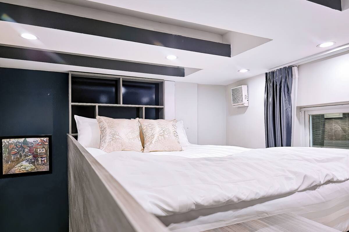 A modern bedroom with a bed, a headboard with built-in shelves, a ceiling light, an air conditioning unit, and a window with curtains.