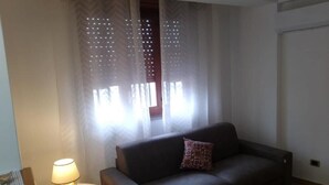 A living room with a dark sofa, a small table with a lamp, and sheer curtains with a pattern.