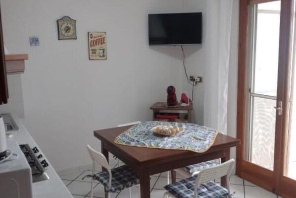 A small kitchen with a microwave, a coffee machine, and a table with a tablecloth. There is a TV mounted on the wall and a door with a glass panel.