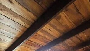 A room with a wooden ceiling, a bed with a wooden frame, a bedside table, and a window with curtains.