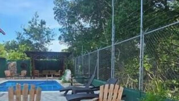 An outdoor seating area with wooden chairs and a table, surrounded by a pool and greenery.