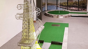 A miniature golf course with a green putting surface, a metal structure resembling a tower, and a backdrop featuring the Eiffel Tower.