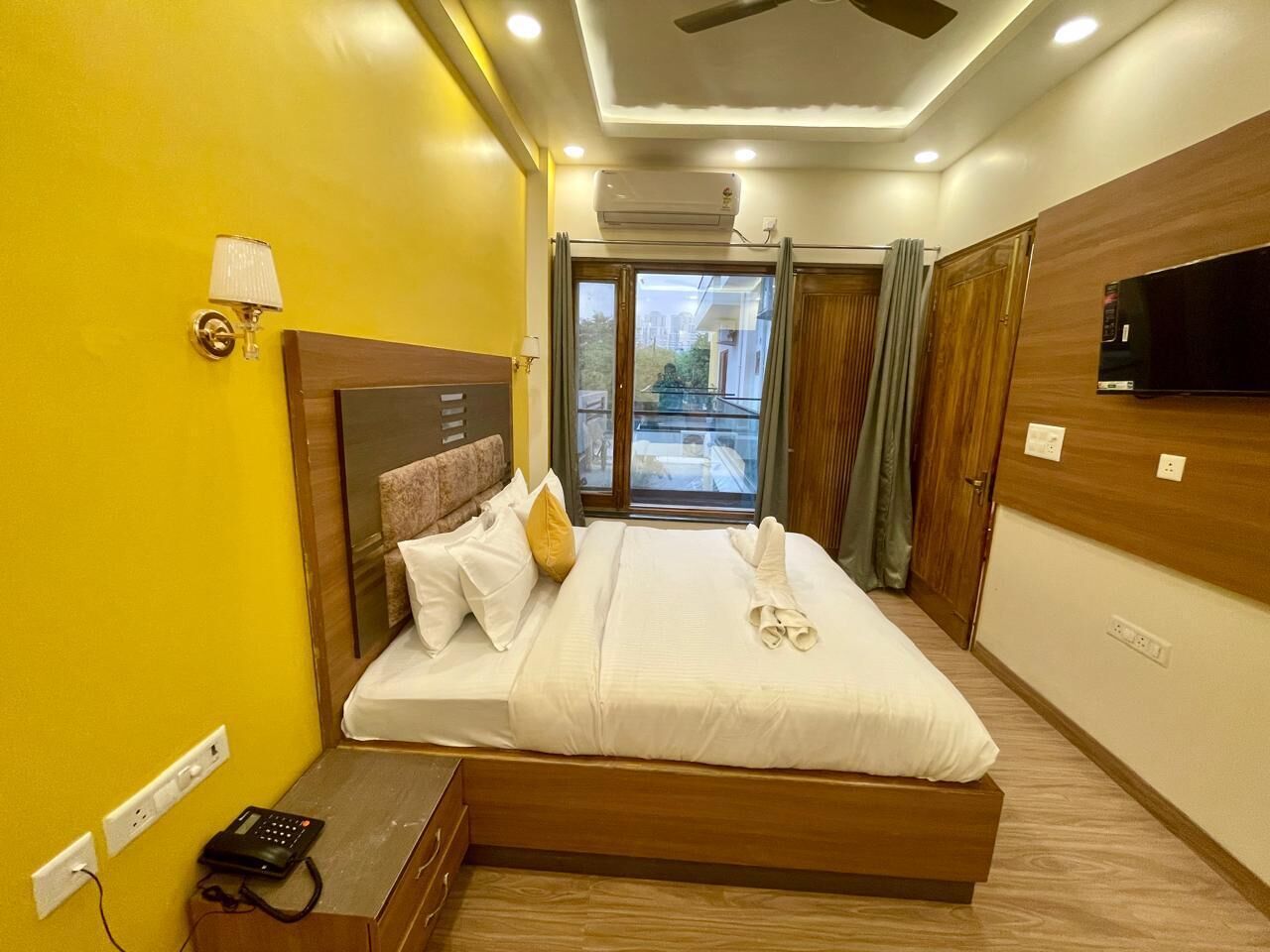 A hotel room with a bed, bedside table, lamp, air conditioning unit, and a television on the wall.