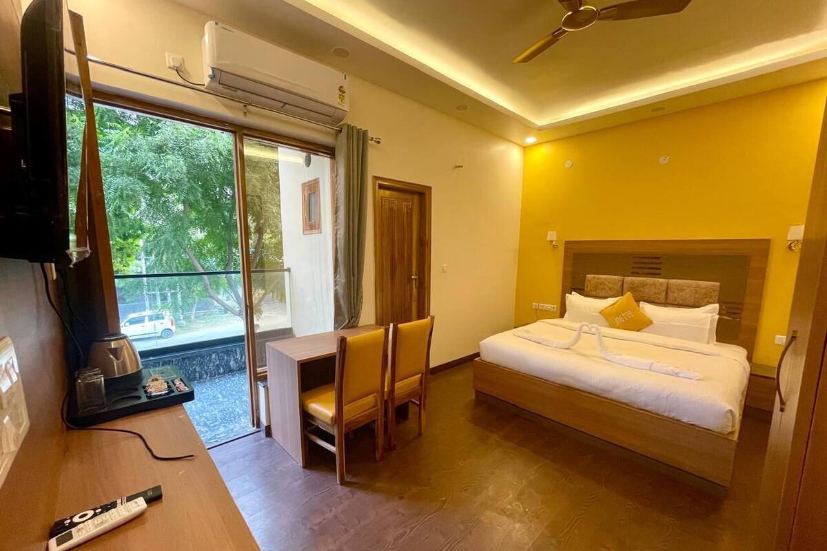 A hotel room with a bed, a desk with a chair, a TV, and a balcony with a view of trees.