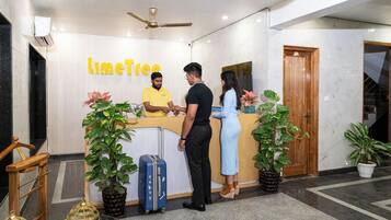 A reception desk with a sign reading "LimeTree," a man in a yellow shirt, a woman in a blue dress, and a man in black. There are potted plants and a blue suitcase.