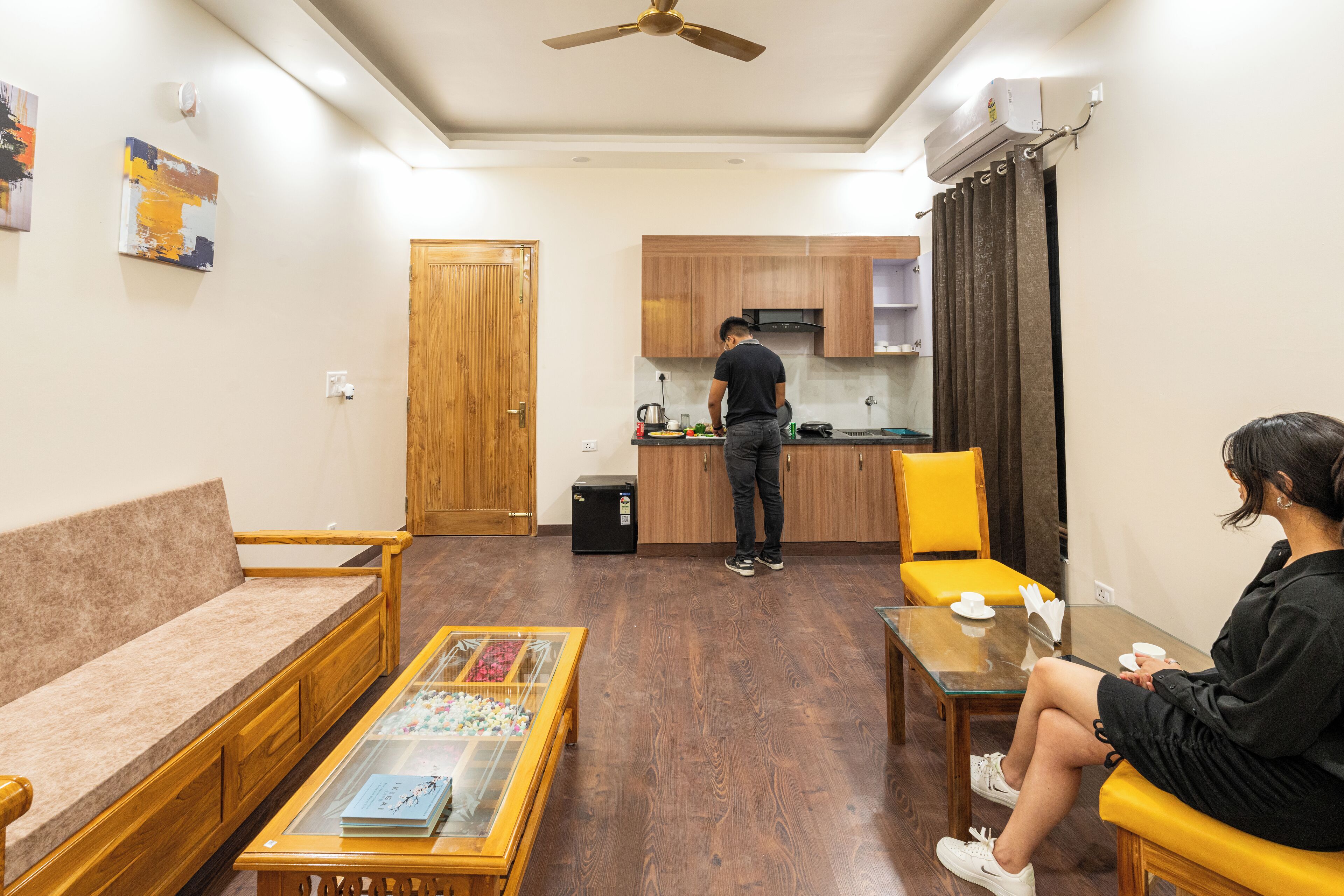 A compact living space with a sofa, a coffee table with a magazine, a dining table with a cup, a kitchen area, and a person cooking.