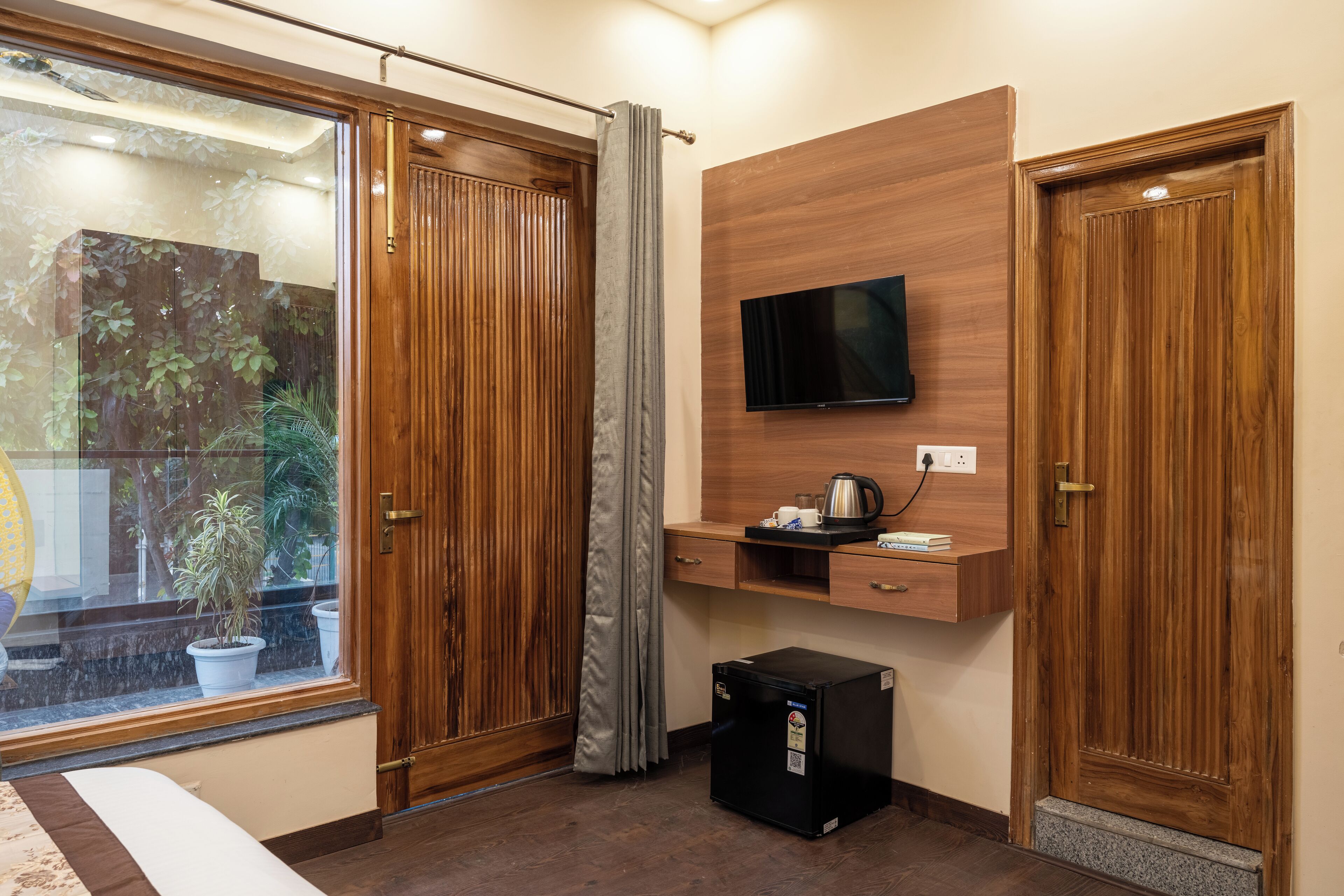 A hotel room with a sliding wooden door, a TV mounted on a wooden panel, a small desk with a kettle, and a refrigerator.