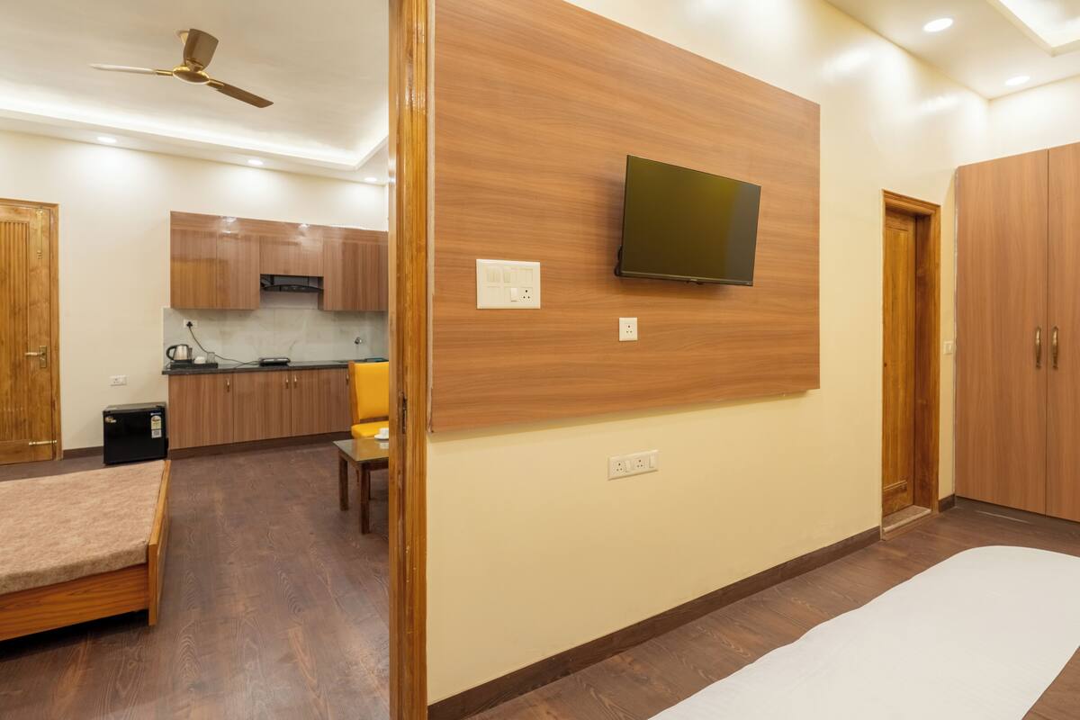 A modern room with a bed, a TV mounted on the wall, wooden cabinets, and a kitchen area in the background.