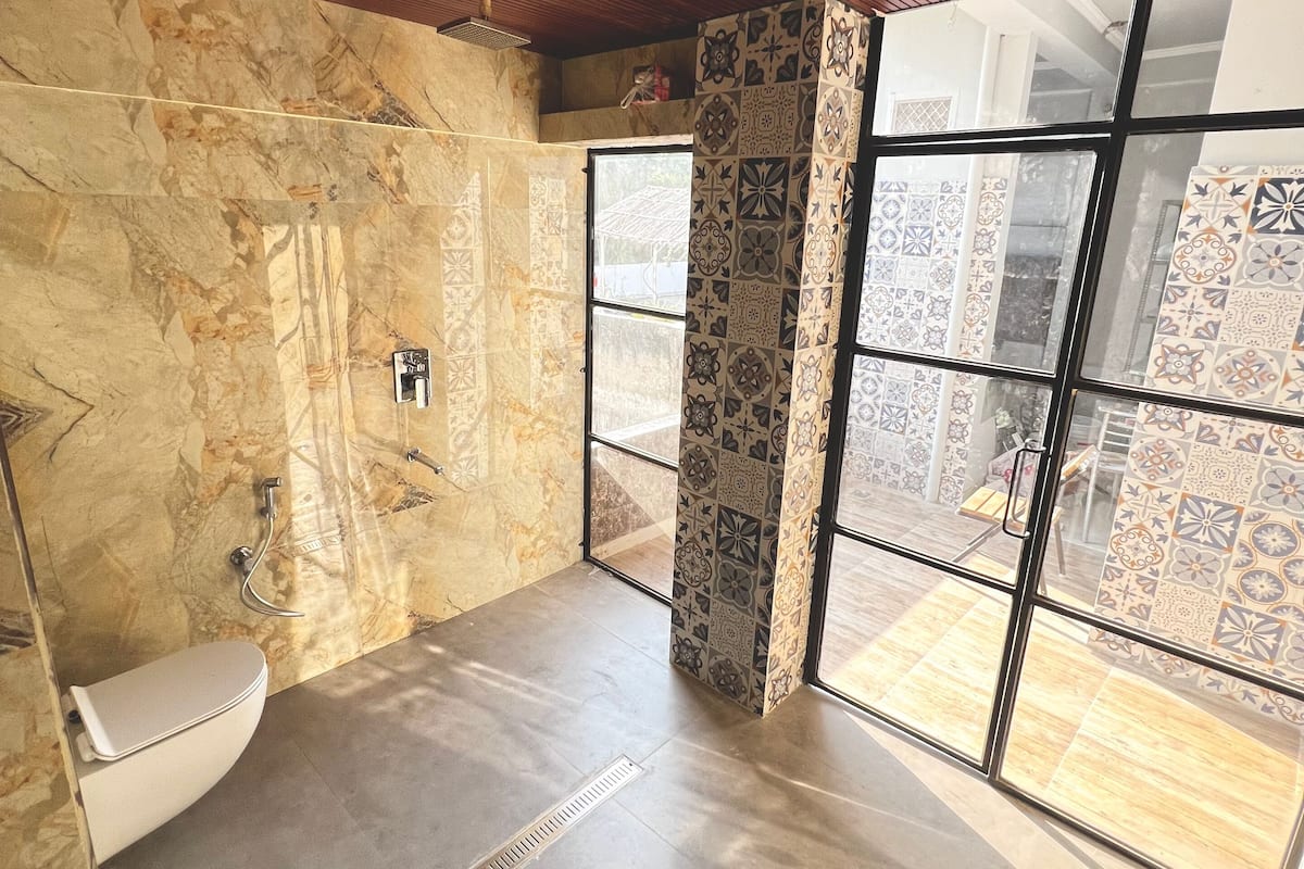 A bathroom with a marble wall, a toilet, a shower, and a large window with patterned tiles.
