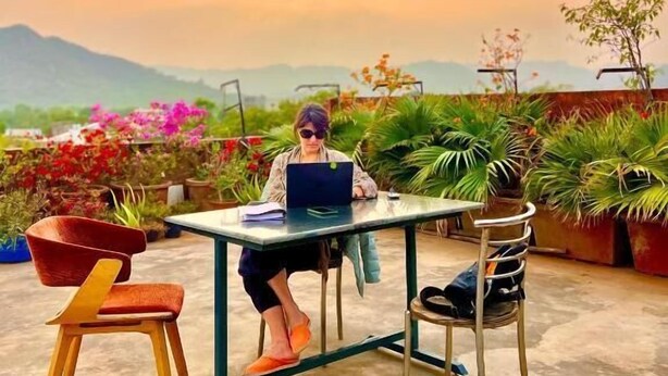 A person working on a laptop at an outdoor table with chairs, surrounded by potted plants and flowers, under a sunset sky. - Bhoora House (Amer)