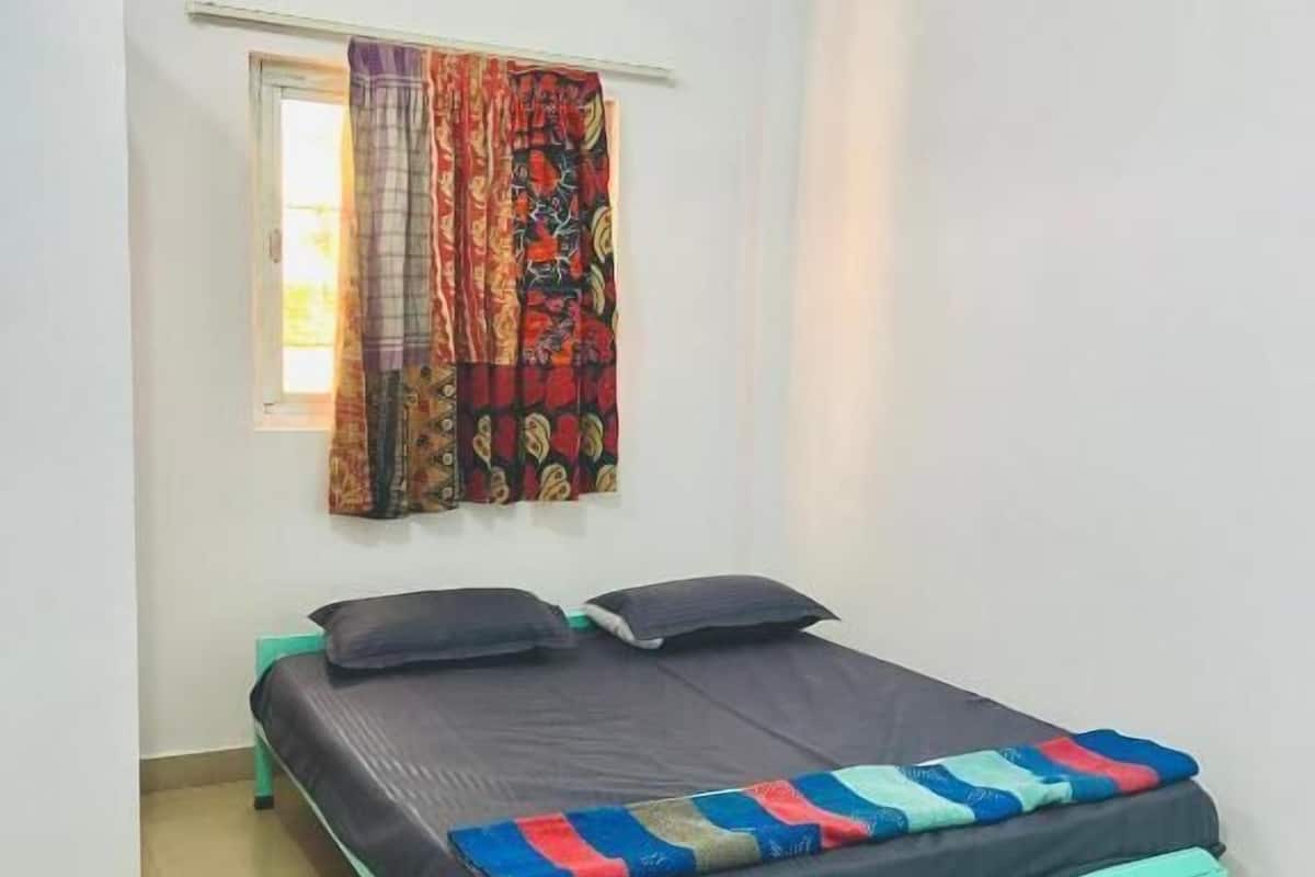A single bed with a teal frame and a colorful quilt, a window with patterned curtains, and a white wall.