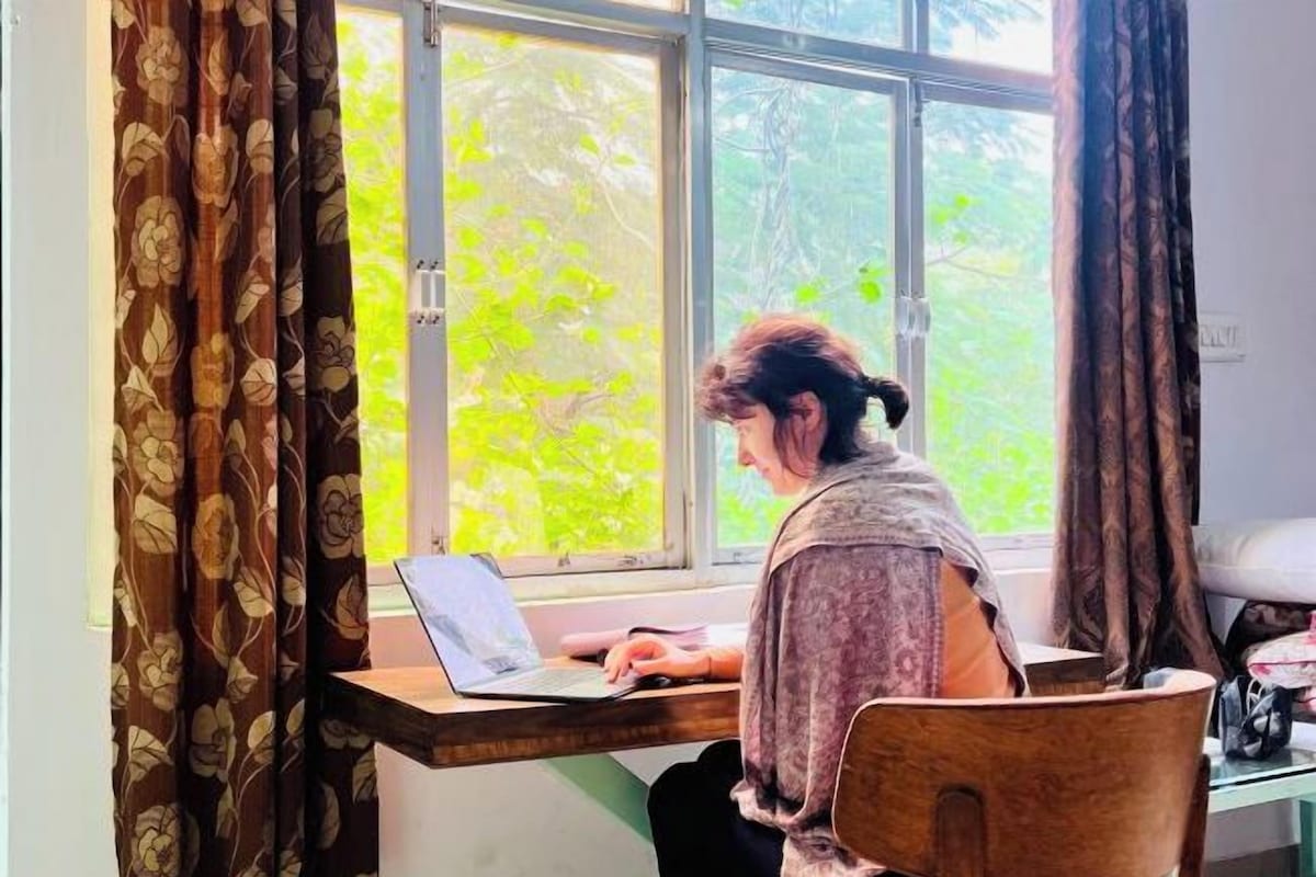 Una persona sentada en un escritorio de madera junto a una ventana con cortinas de flores, usando un ordenador portátil.