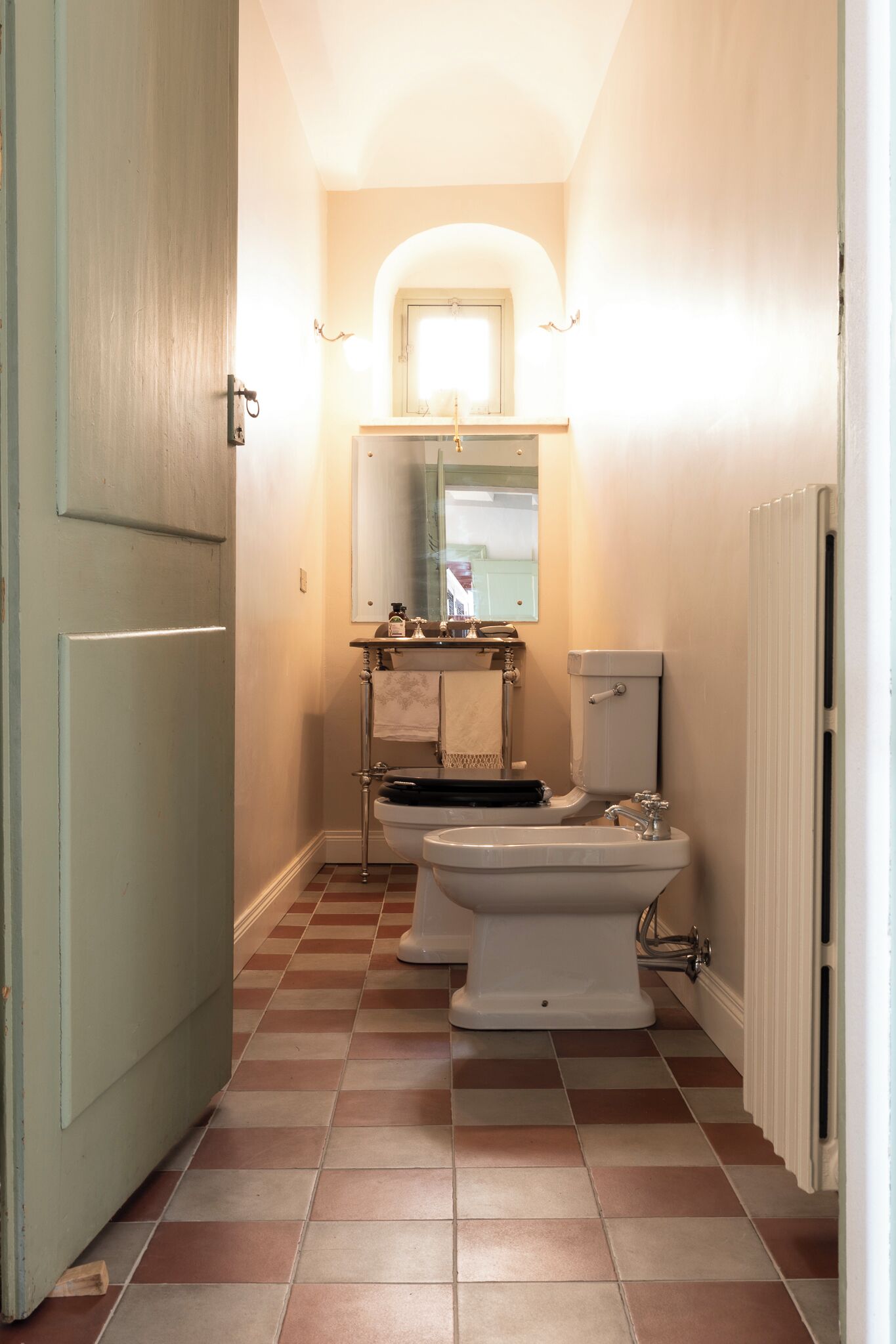 A narrow bathroom with a toilet, bidet, and sink, tiled floor, and a door on the left.