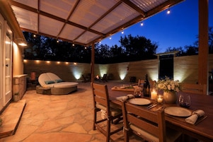 An outdoor patio with a covered seating area, a dining table set for two, and a fire pit in the background.