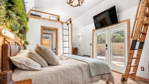 Ein Schlafzimmer mit einem Bett, einem Fernseher, einer Leiter und Blick durch eine Glastür.