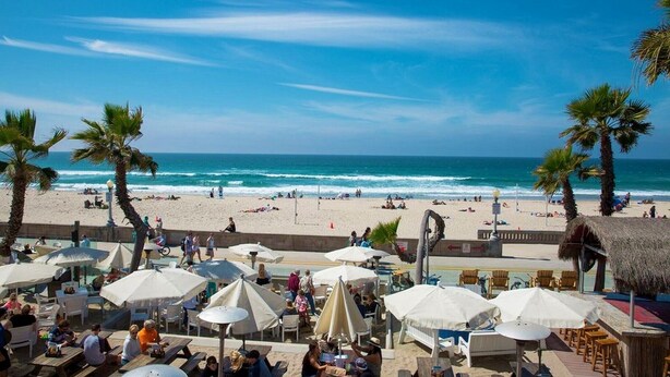 Um café à beira-mar com mesas ao ar livre e guarda-sóis, com vista para uma praia de areia e o oceano.