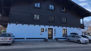 Un edificio de dos plantas con fachada de madera oscura y una sección inferior azul claro. Se observan varias ventanas con contraventanas, una puerta con un pequeño porche y una escalera visible que conduce a la entrada.